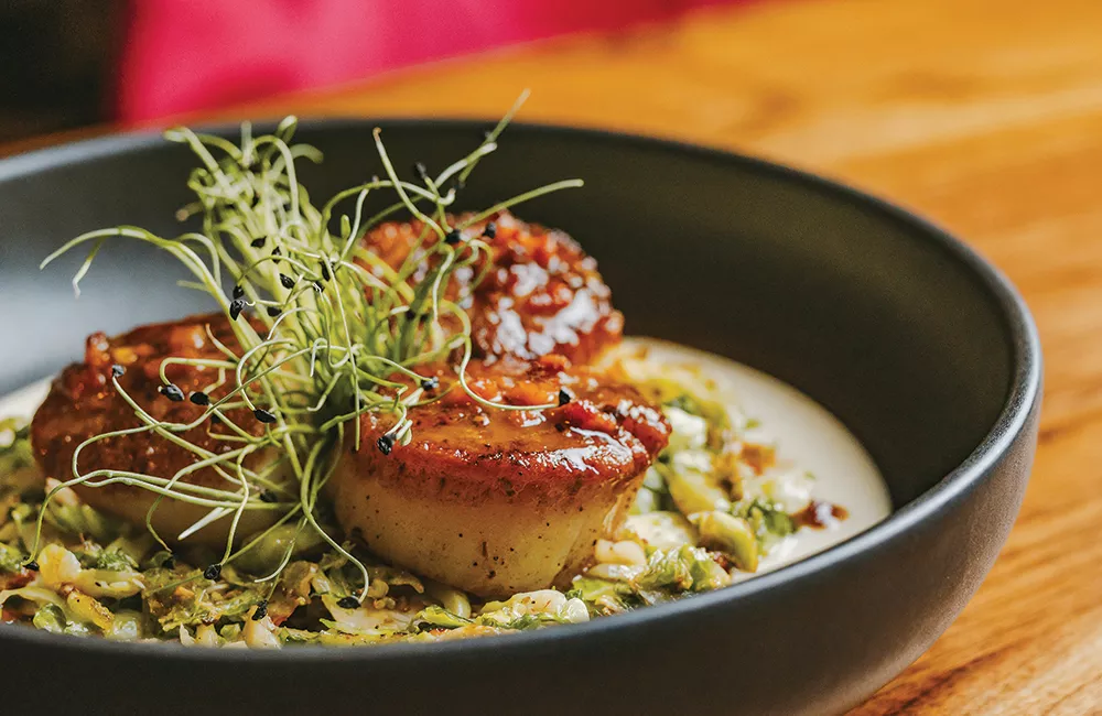 Seared scallops topped with greens on a dark plate at Dear Irene