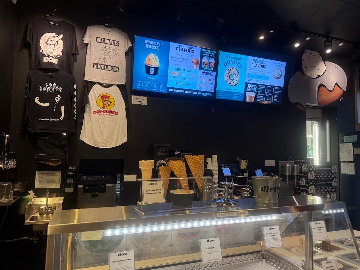 Doe Donuts’ interior counter with a glass pastry case, waffle cones, illuminated menus, and hanging shirts.