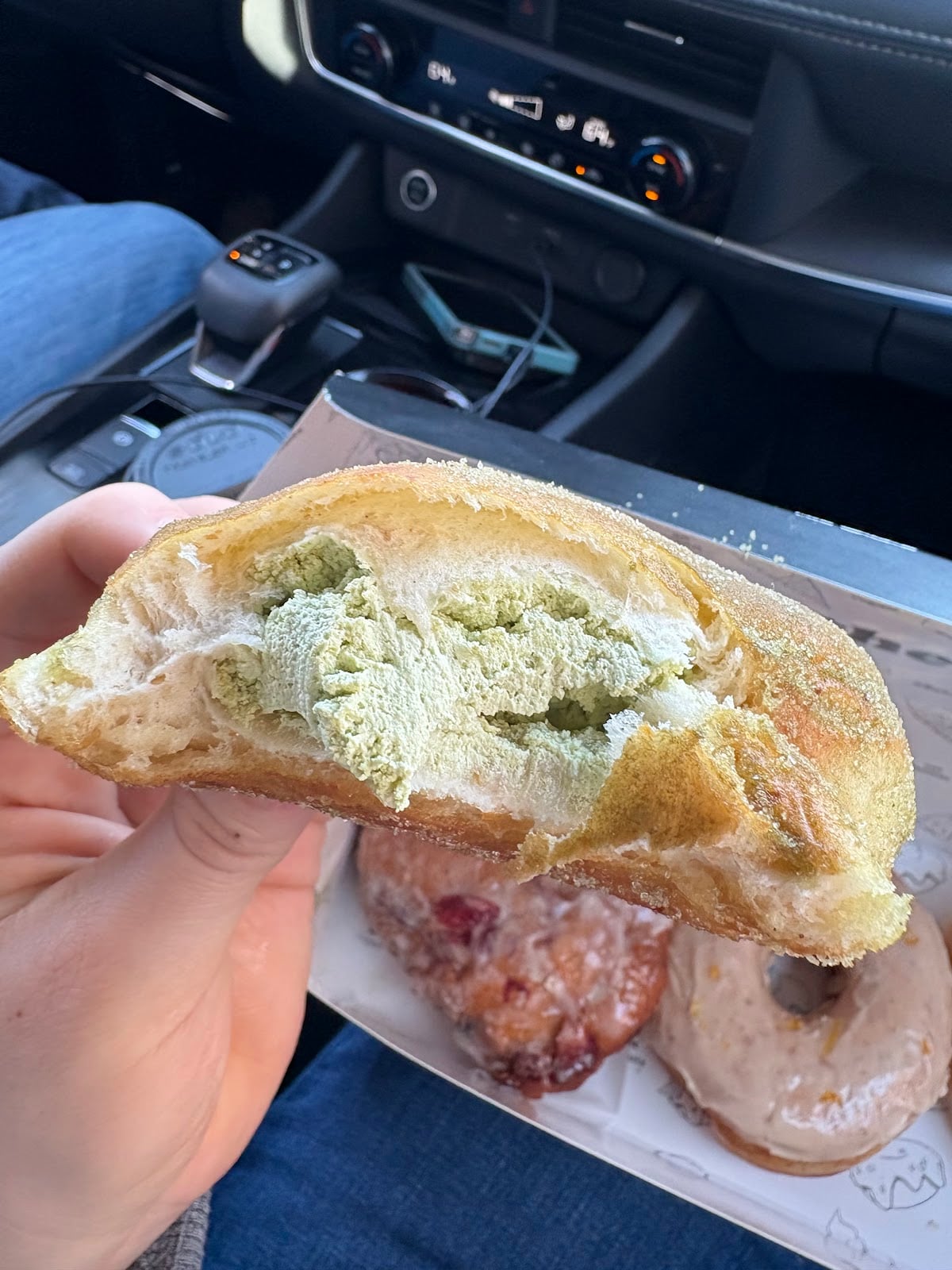 A cut-open sugar-coated donut revealing pale green matcha filling, with two other donuts in the box below.