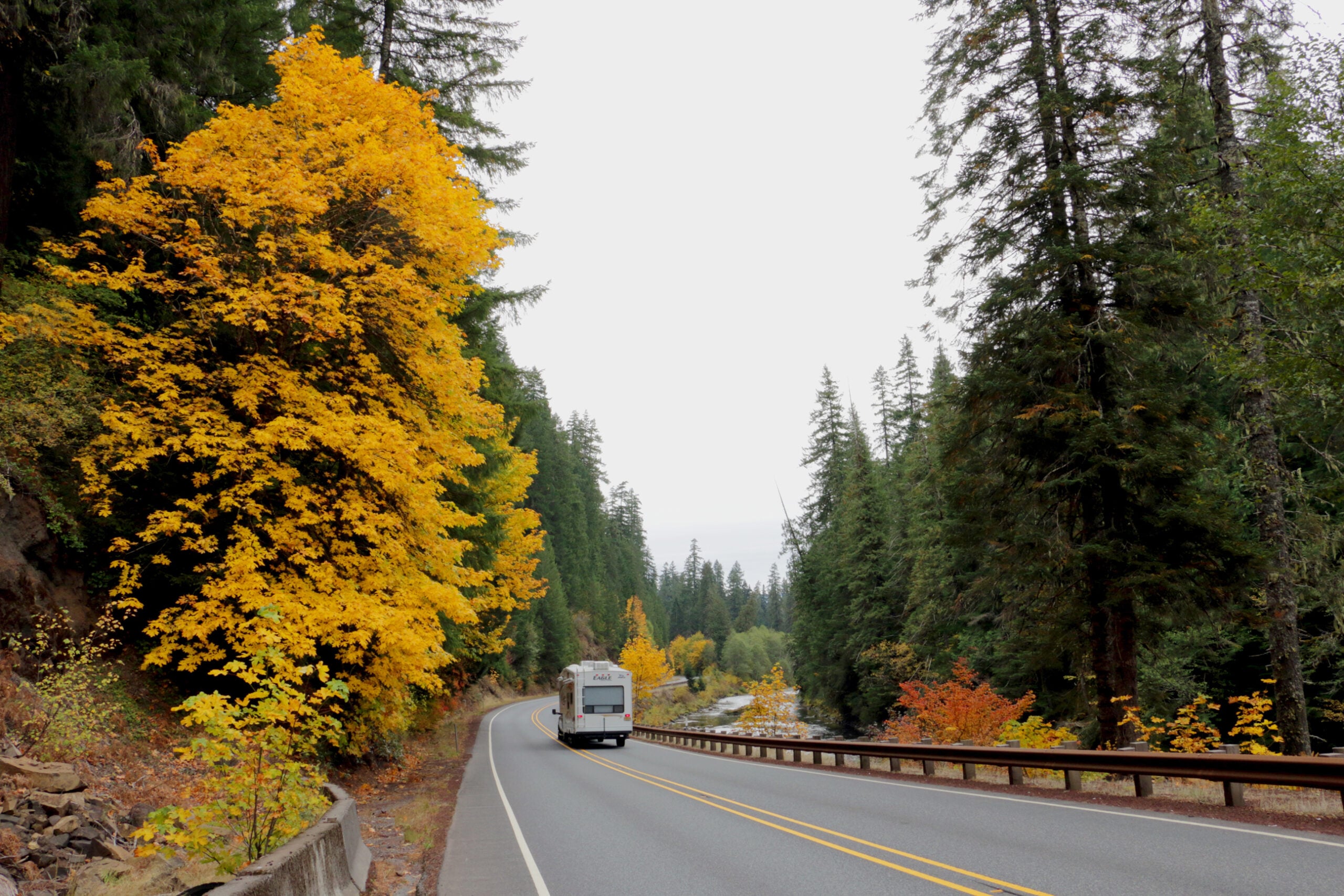 An RV travels along a curving two-lane highway beside a river and forest with yellow autumn leaves.