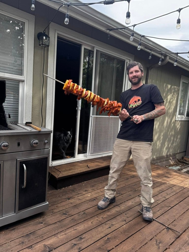 Kaylub Giamanco stands on a wooden deck holding a large skewer of meat and pineapple.