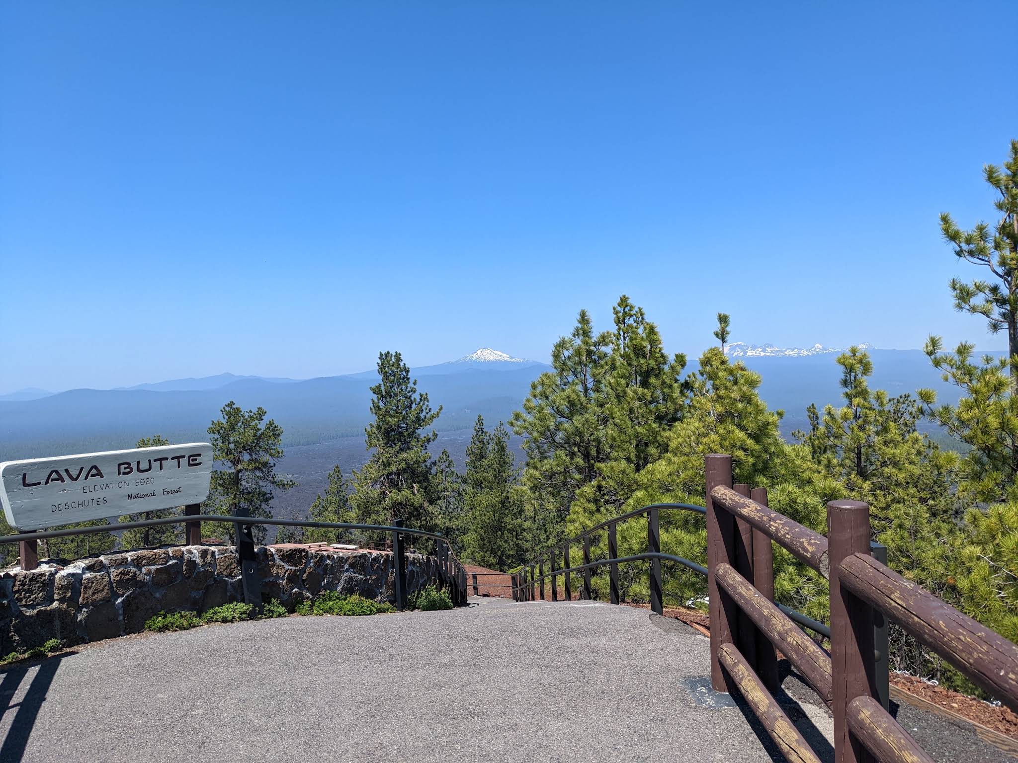 Lava Butte summit sign beside a paved overlook with pine trees and Cascade peaks