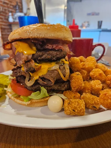 A stacked burger with bacon, melted cheese, onions, lettuce, and tomato beside golden tots
