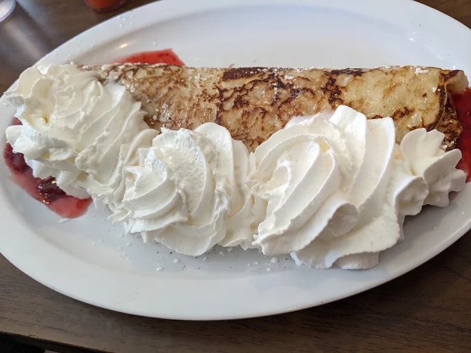 A folded golden brown breakfast dish with red fruit sauce and whipped cream on a white plate