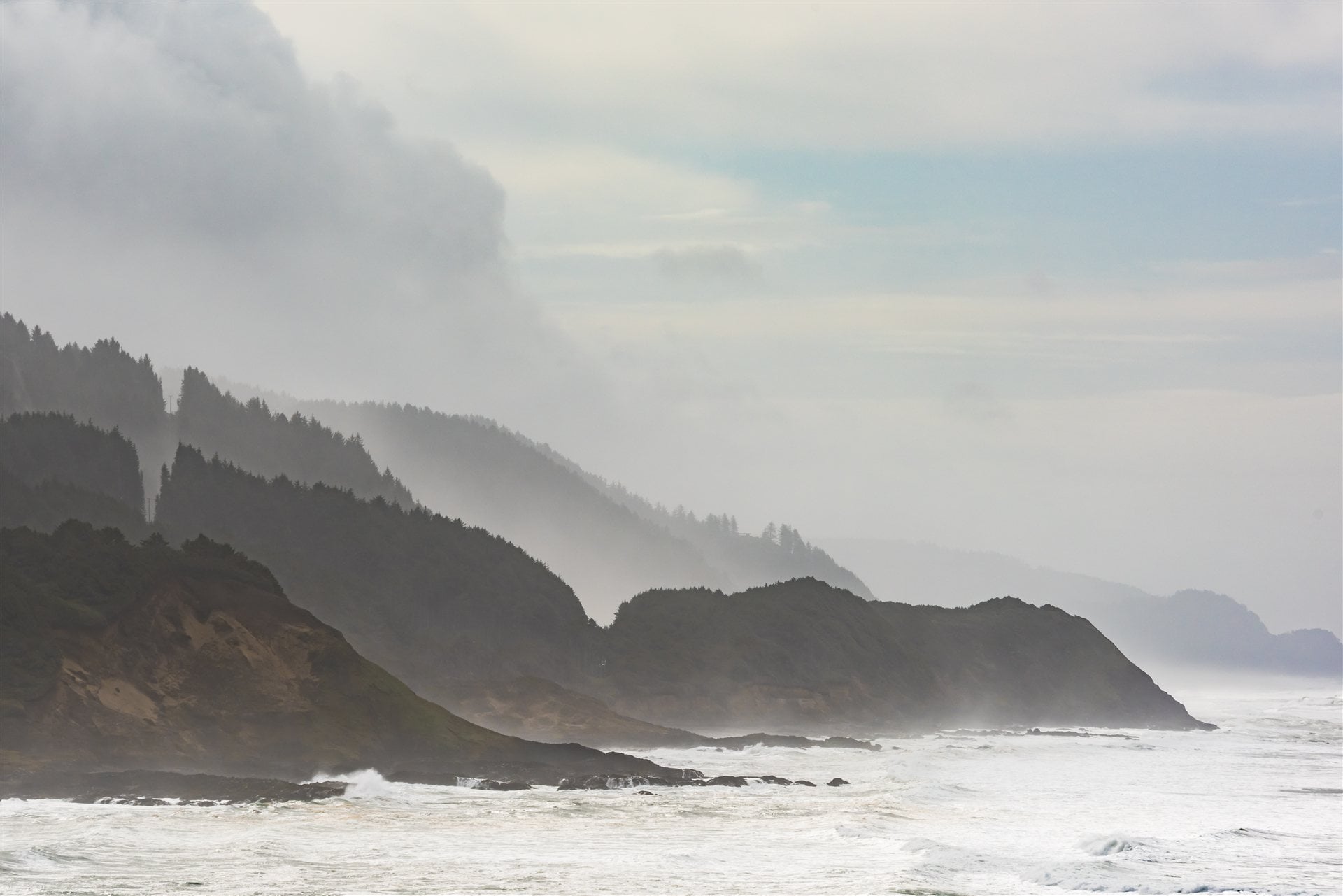 Misty forested cliffs above waves along the Oregon Coast under a stormy sky.