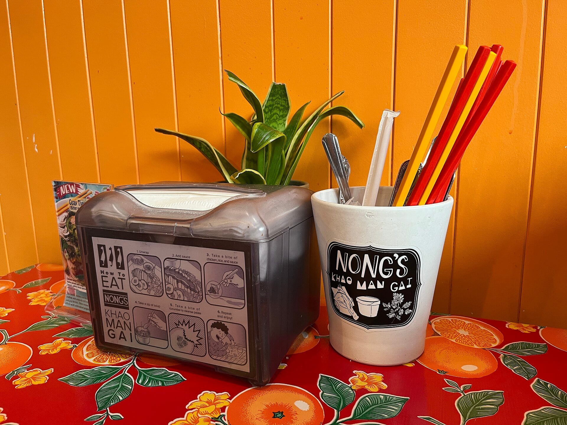 Red and yellow chopsticks beside a small plant and illustrated napkin dispenser on a floral table