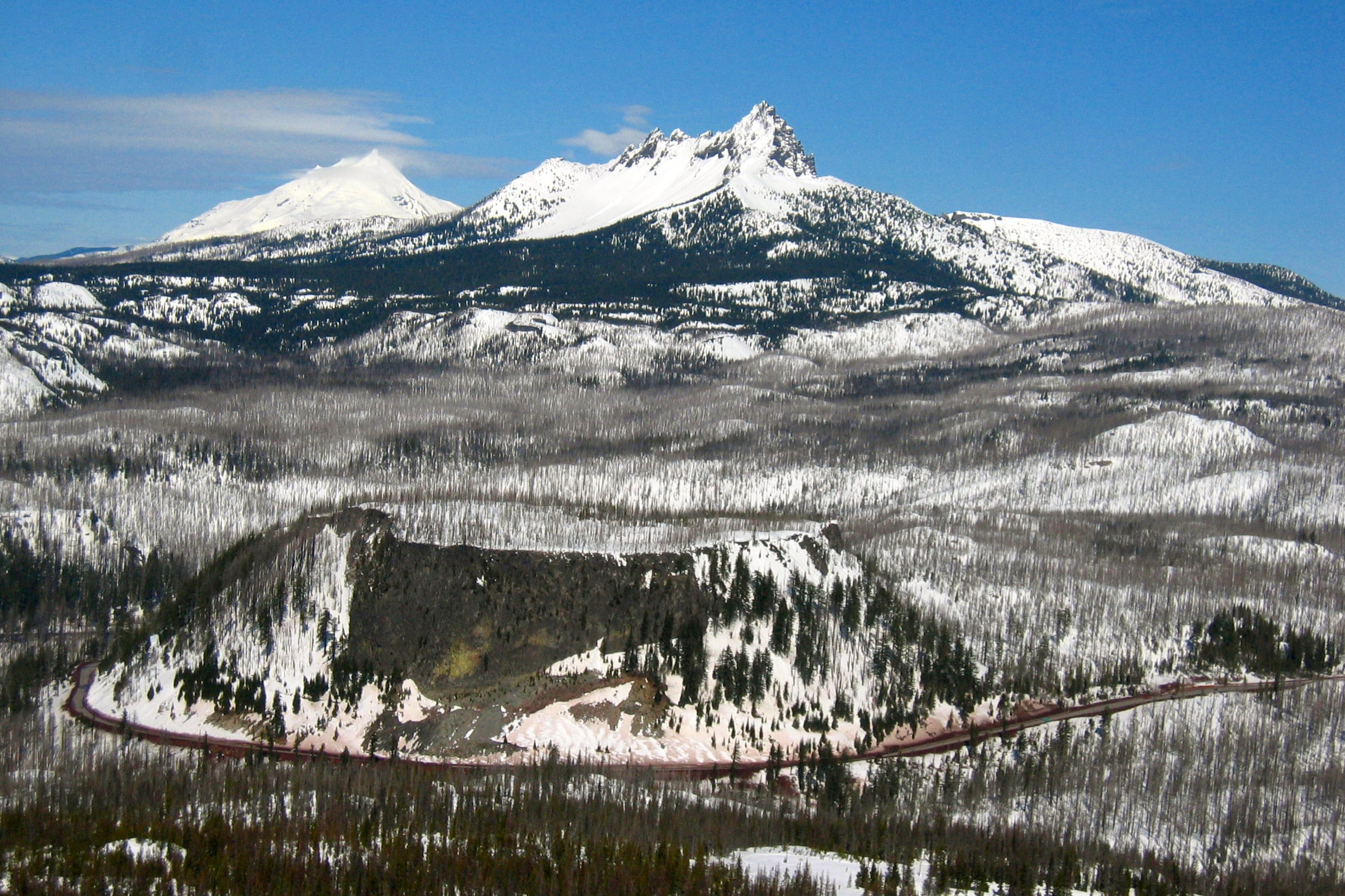 Highway 20 curves below snow-covered Three Fingered Jack, with Mount Jefferson visible to the left.