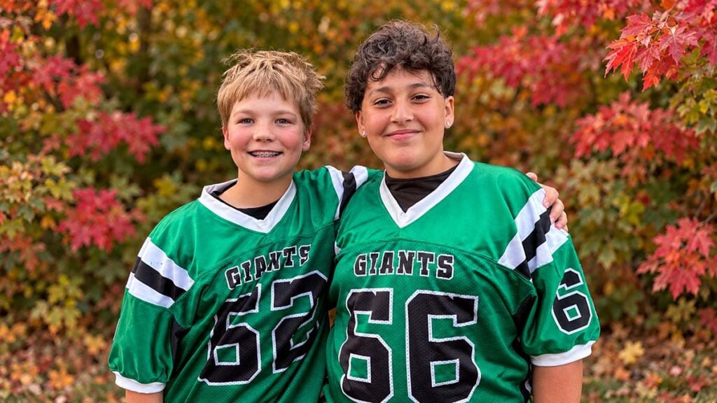 Two Bend students smile with their arms around each other, wearing green Giants football jerseys in front of colorful autumn leaves.