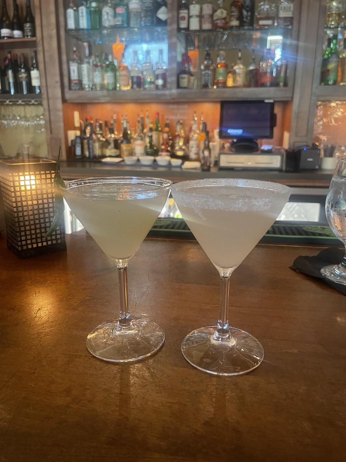 Two pale cocktails in stemmed glasses on the wooden bar at Vault 244, with bottle shelves behind them.