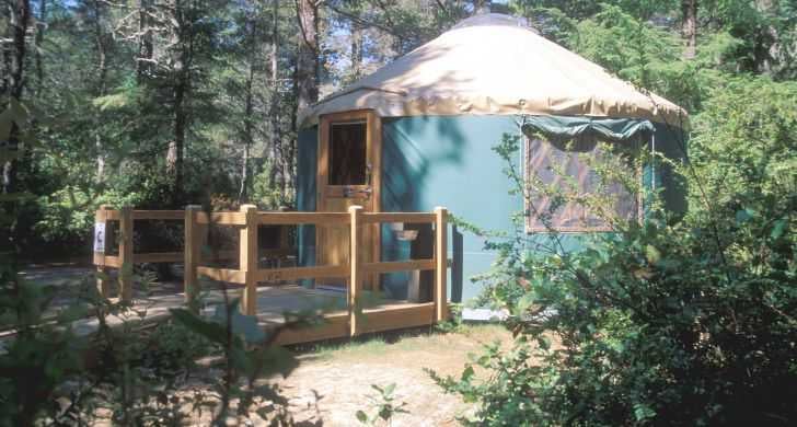 Green yurt with a tan roof and wooden entry deck among trees at William M. Tugman State Park.